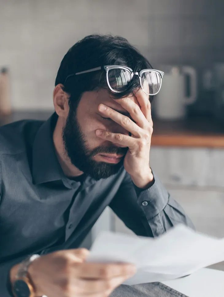 A bearded man with eyes shut holding his face in his hands.