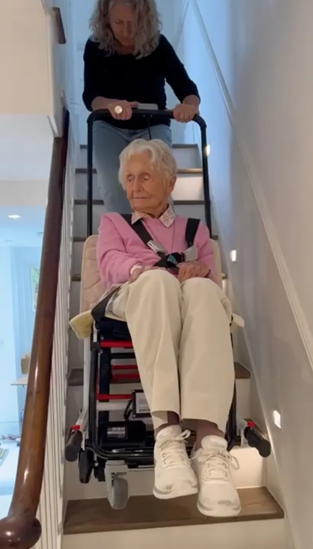 A woman pulls an old lady seated in a mobile stairlift up a narrow wooden staircase.