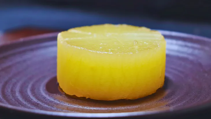 A beautifully braised daikon radish served on a ceramic plate, with its translucent yellow hue showcasing the deep absorption of dashi broth, representing a classic Japanese dish.