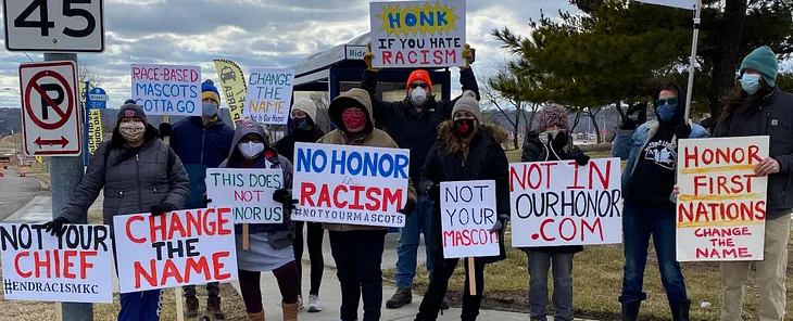 A group of ten people wearing winter coats, masks, and hats standing on the side of the road. Each is holding a handwritten sign, which read: “Not your chief #endracismkc,” “Change the name,” “Race-based mascots gotta go,” “This does not honor us,” “Change the name #not in our honor,” “No honor in racism #notyourmascots,” “Honk if you hate racism,” “Not your mascot,” “Not in our honor .com,” and “Honor first nations change the name.”