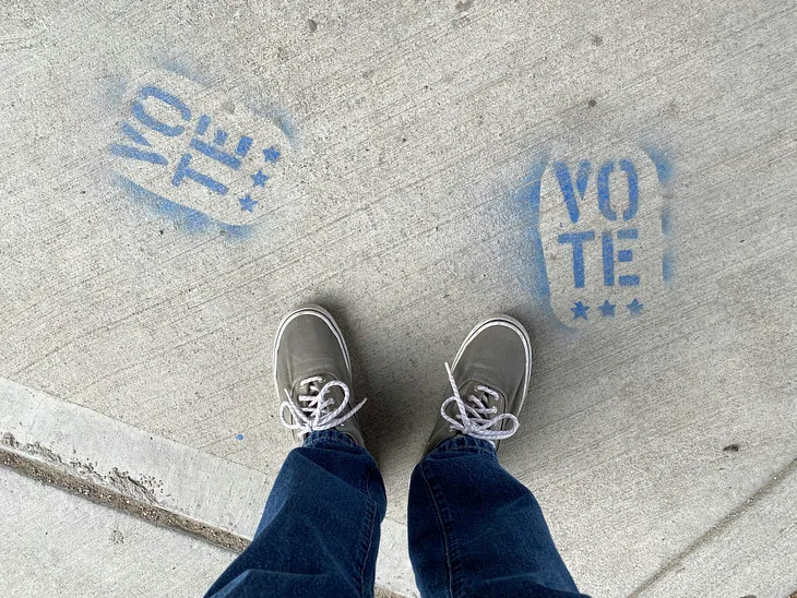 VOTE! pavement art. Person in blue denim jeans and white sneakers standing on grey concrete floor.