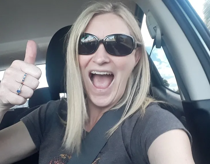 Photo of Author giving the thumbs up from a selfie taken inside the car, wearing sunglasses and a grey t-shirt and with blonde hair just past her shoulders.