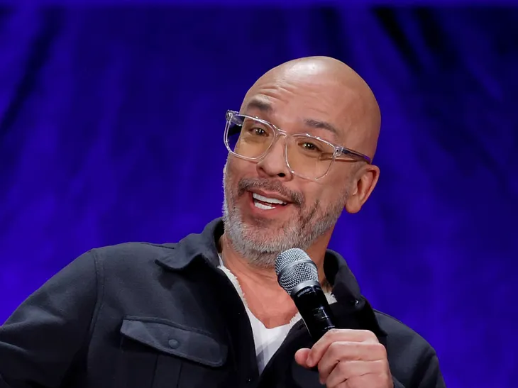 Image of comedian Jo Koy in front of a blue wall with a microphone.