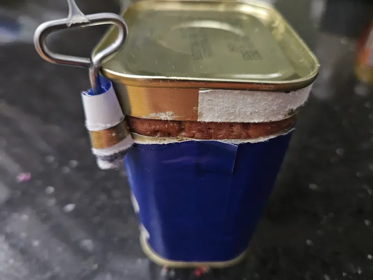 Blue paper-wrapped, cuboid stainless steel tin. A glimpse of corned beef is visible in the thin gap revealed by the jagged track of the tin’s minimal wire key. The key is wrapped in the paper it has torn from the tin’s label.