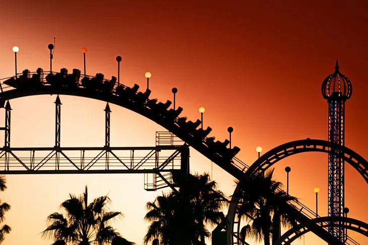 A silhouetted roller coaster against a vibrant sunset, framed by palm trees and illuminated ride elements.