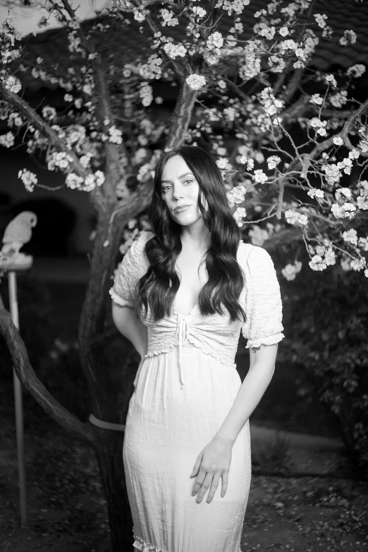 Girl in long white dress stands in front of a blossoming fruit tree in garden. Black and white photo.