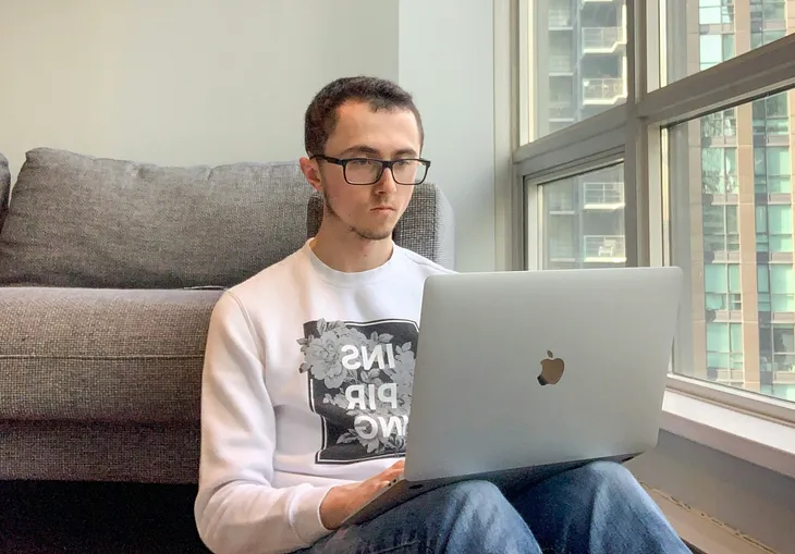 A man in his mid 20s wearing a white sweather and blue jeans. He’s sitting on the floor against a couch. He’s resting a MacBook against his lap. He has black hair and glassses. On the right-hand side of the frame is a window.