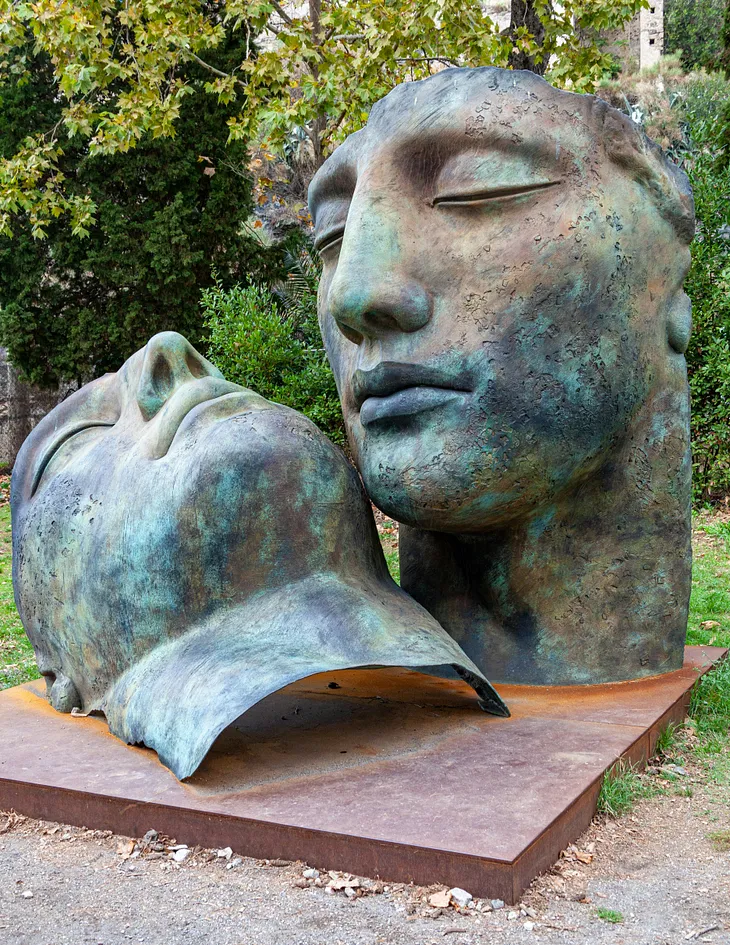 Sculpture of two gray heads near green trees during daytime.