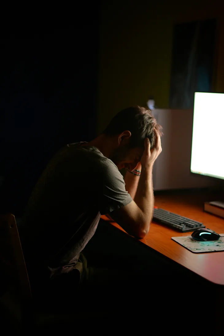 A man sits in the dark with his face buried in his hands. The only light comes from his computer screen, giving the vibe of endless struggle