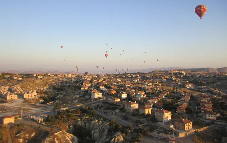 Many hot air balloons flying low over a city at sunrise