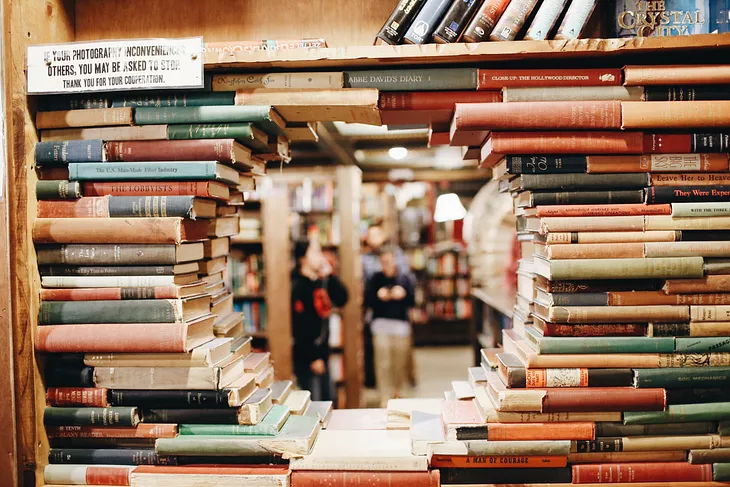 A library shelf on which books of various sizes and colors have been arranged so that they form the boundaries of a circle in the middle of the shelf. Through the circle, the people on the other side of the bookshelf can be seen (out of focus). On the top left of the shelf, there is also a sign about taking photographs.