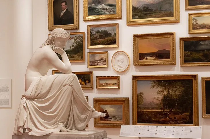 A neoclassical sculpture of a seated woman sits in front of a salon wall of paintings.