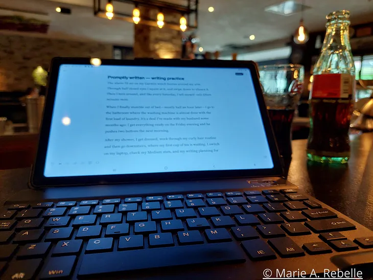 Image shows a tablet, the screen filled with text, and a keyboard in front of it. To the right of the tablet is a bottle of cold drink and in the background you see a restaurant.