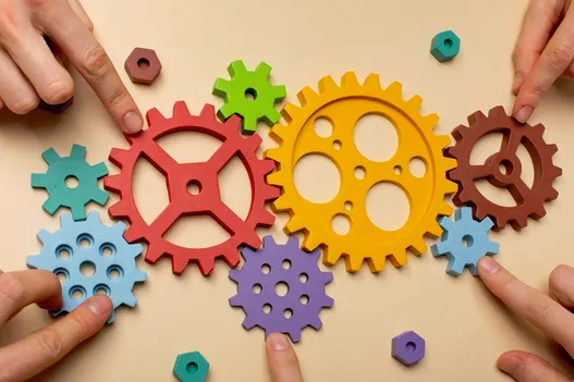A figure with several people’s hands pushing together assorted mechanical gears.