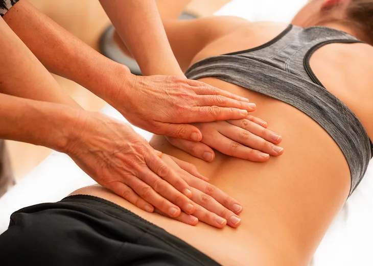 Photo of a woman’s torso as she lies on her back. Other people have their hands on her back like they’re giving her a massage.