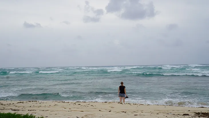 A woman looks out at a stormy ocean from the beach with shoes in her hand