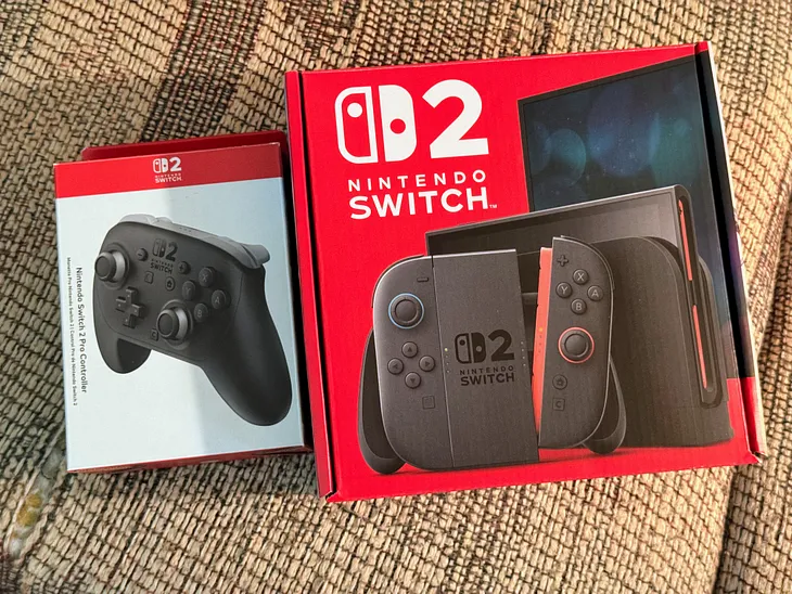 A Switch 2 Pro Controller Box (left) and a Switch 2 Box (right) sitting next to each other on my slightly ugly patterned couch.