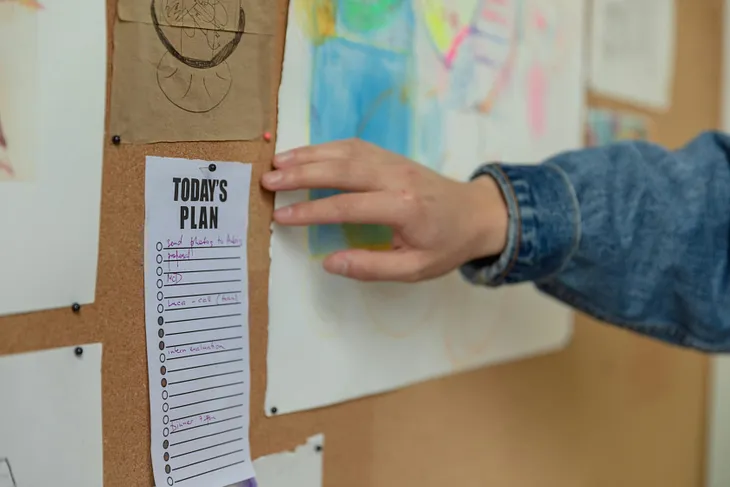 A hand reaching for a bulletin board containing a list headed with Today’s Plan.