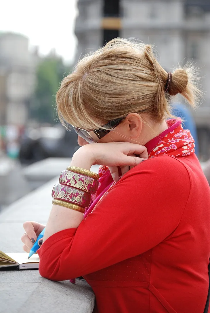 Blonde woman, hair tied back wears sunglasses, bangles and red clothes, looks thoughtful and writes in a journal