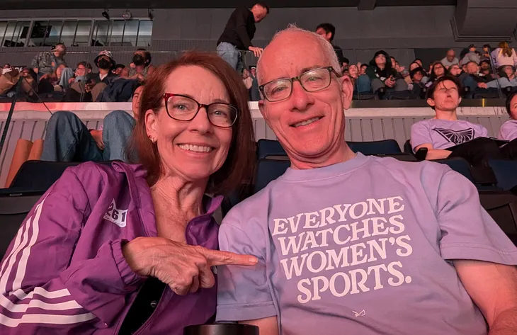 A photo of the author and her husband at the Golden State Valkyries game. She points to his violet shirt that says “Everyone Watches Women’s Sports.”