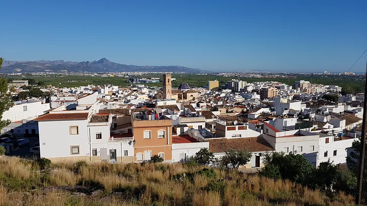 A view from a hill looking over the white washed houses of Oliva, a small Spanish town. There are gree luch orange groves in the diastance beyond the houses.