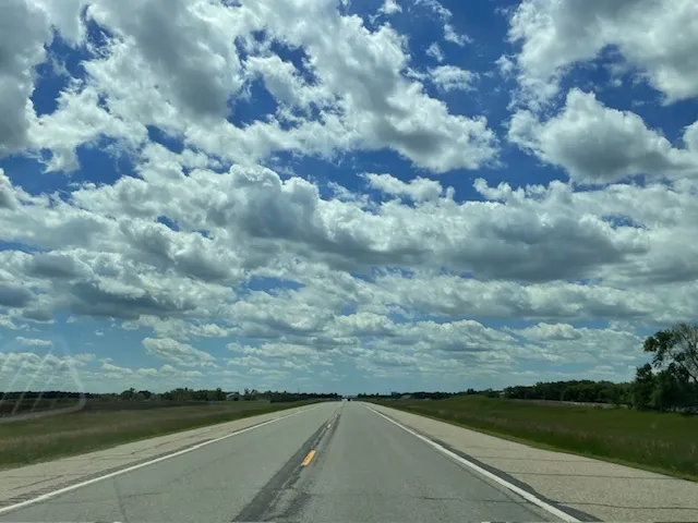 Color photo of open landscape, paved highway, and cloud-filled sky