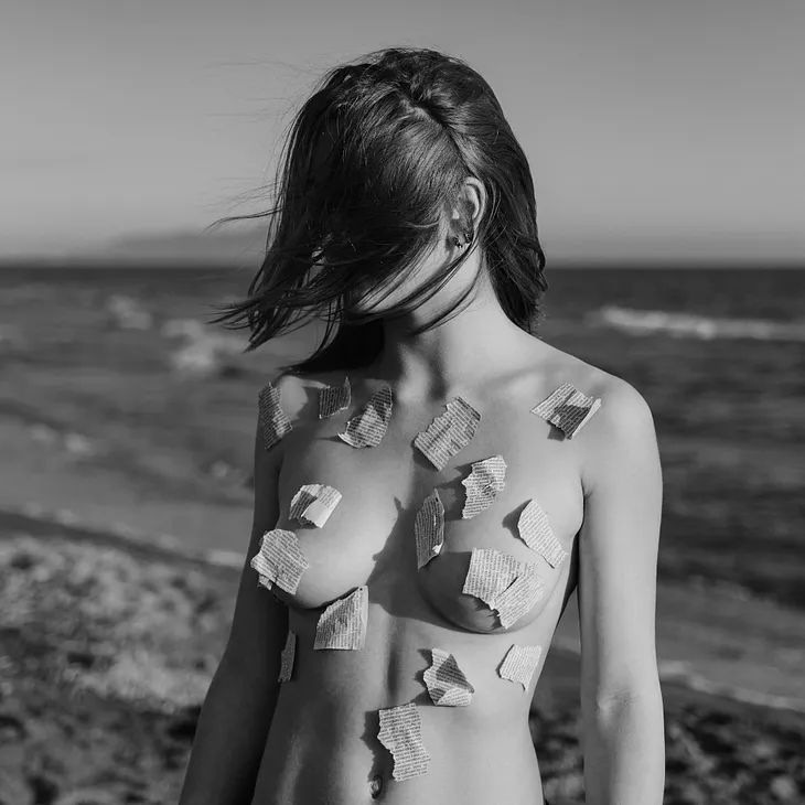 Black and white image of a nude woman on the beach with scraps of paper hiding her nudity.