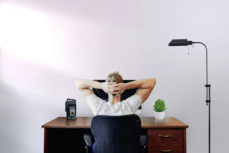Man sitting at his computer with hands crossed behind his head.
