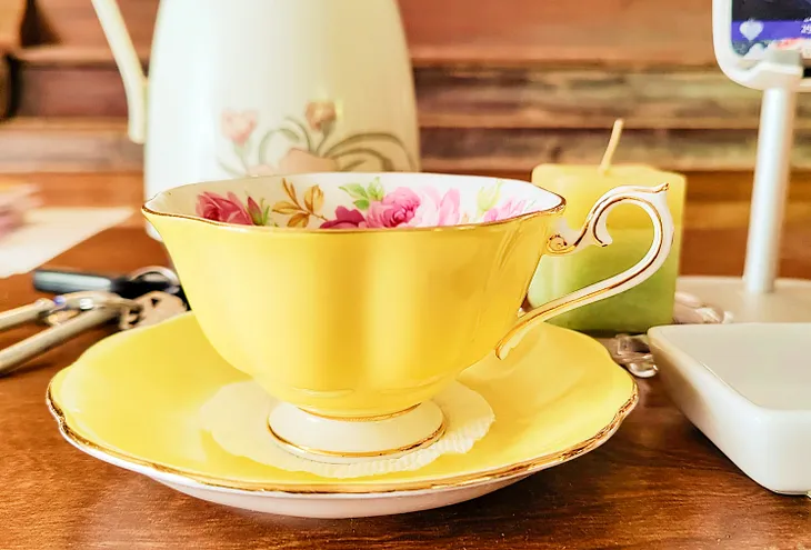 Scalloped and gilded-edge yellow bone China teacup with pink cabbage roses and green leaves painted on the inside of the cup. It sits in its matching yellow saucer and holds golden chamomile tea.