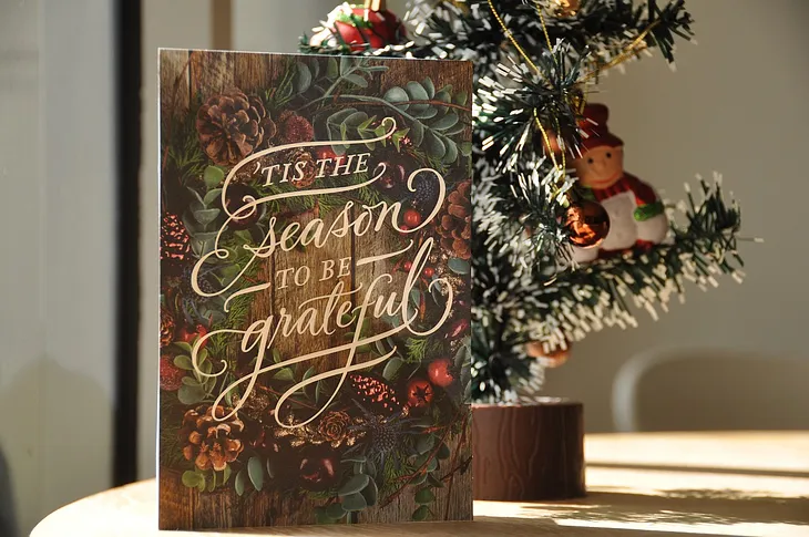 A book standing next to a christmas tree and the civer states, ’Tis the seasin to be grateful’.