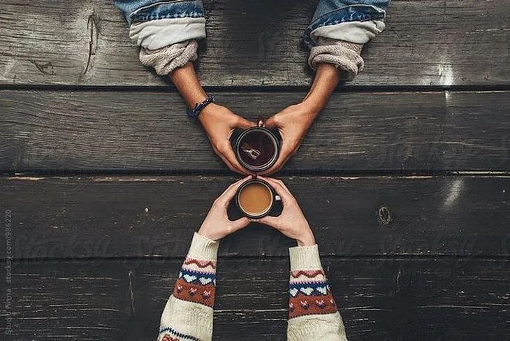 Two hands holding cups filled with Coffee/Tea upon a wooden table.