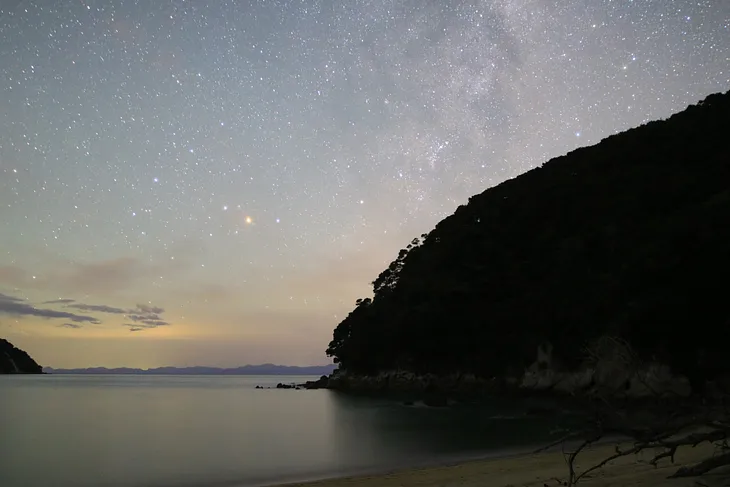 A starry sky above the ocean with a dark mountain on the side.