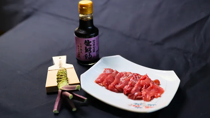 A neatly arranged plate of maguro (tuna) sashimi sits beside a bottle of nama shoyu (raw soy sauce) and a fresh wasabi root with a grater, all placed on a dark background.