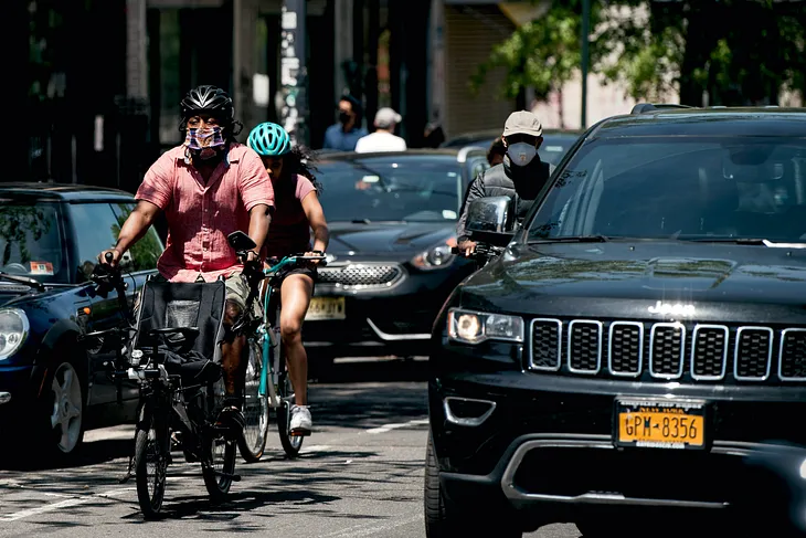 Three cyclists, one with a bike meant to carry a wheelchair-bound rider in the front, pass between a row of parked cars and moving cars. A large SUV is in the foreground.