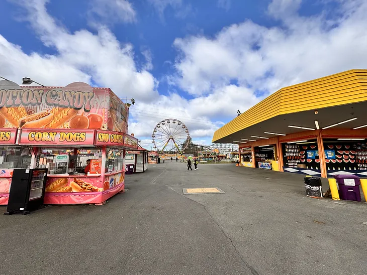 wide angle picture of a stall selling fair food, another stall on the right, and the ferris wheel in the background