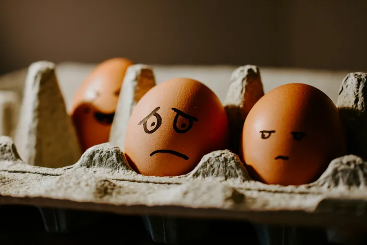 Picture of a sad looking egg in a carton with a neighboring egg watching cautiously.