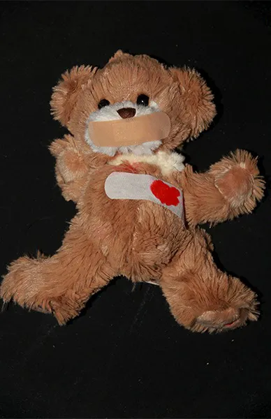 A teddybear with a band aid covering their mouth and a band aid seemingly with blood on it, covering their heart.