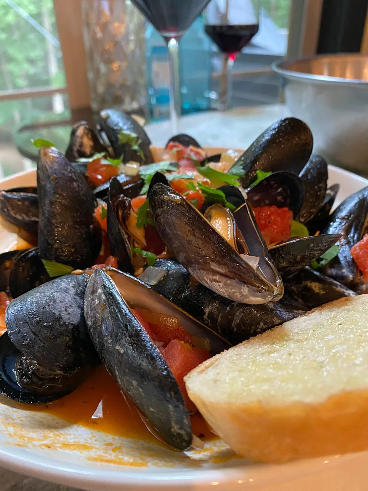 An explosion of visual stimulus with blue-black mussels, red tomatoes, orange mantle, white onion & garlic and topped with forest green flat Italian parsley.