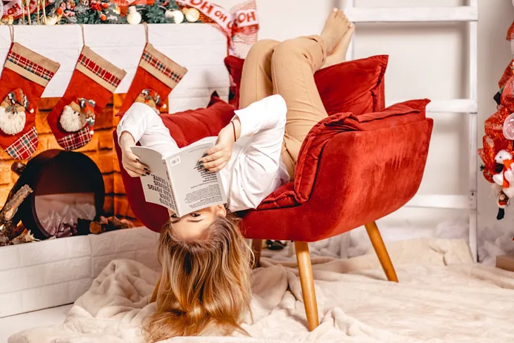 A woman is hanging upside down in a red plush chair with her legs over the back, a book covering her nose, and her blond hair touching the floor. She is in front of a fireplace decorated for Christmas and stockings hung all in a row.