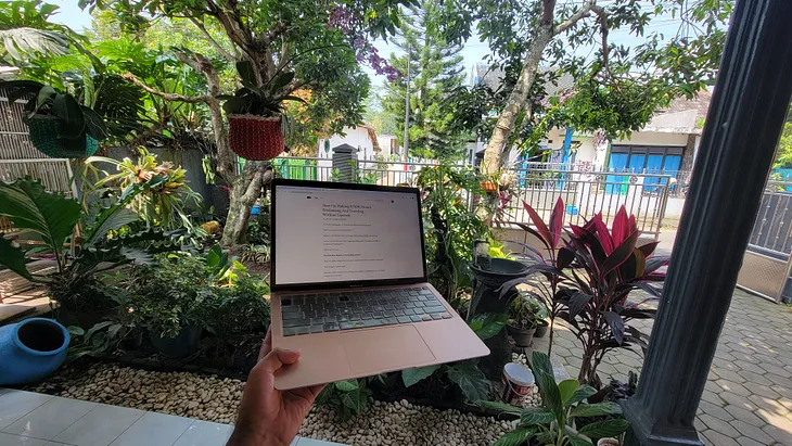 Guy holding his macbook with his left hand in front of greenery.