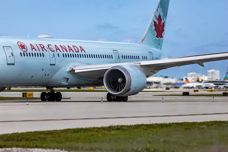 A plane painted in light blue with the red “Air Canada” lettering on the side is parked at an airport tarmac.