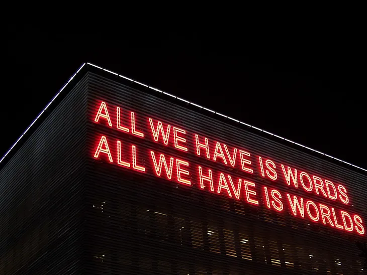 A striking neon sign displays the thought-provoking phrase: “All we have is words, all we have is worlds.”