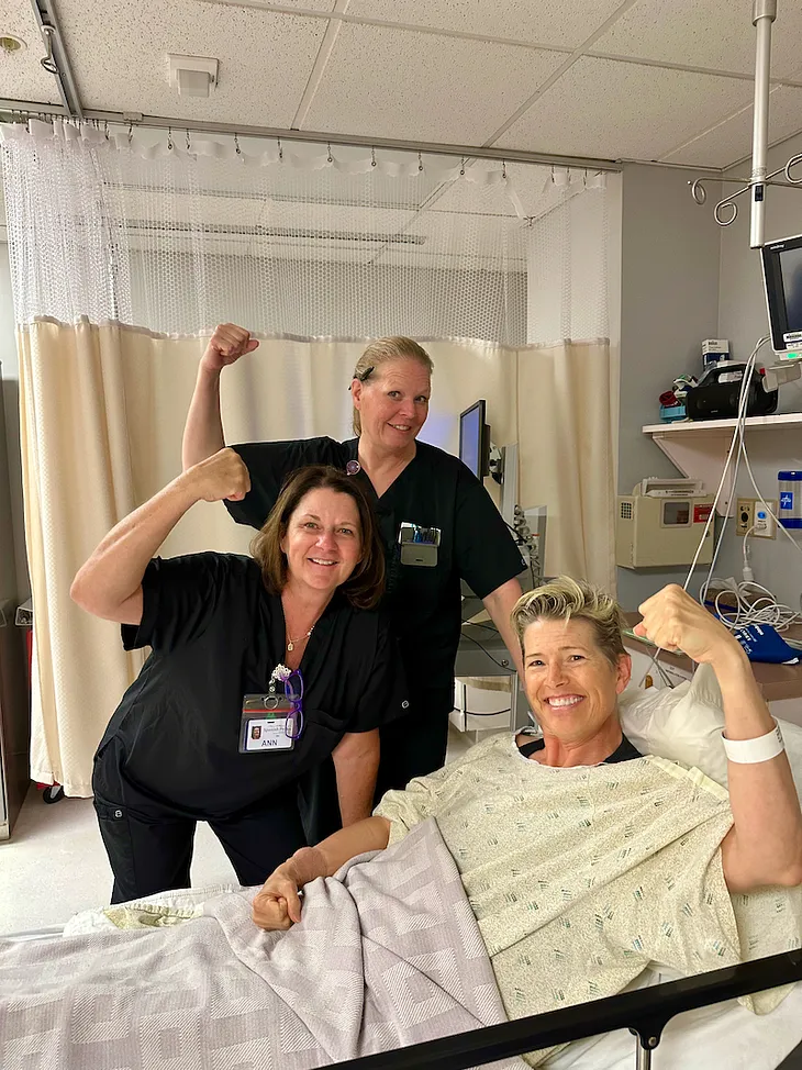 CC in a hospital gown on a gurney posing with two of the surgery staff from Spanish Peaks Regional Health Center each doing a strong arm selfie celebrating her clean and clear colonoscopy!