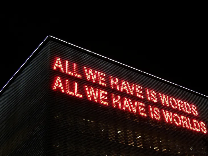 Neon lights on a building forming a sign: “All we have is words, All we have is worlds.”