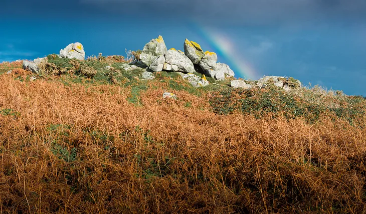 Landscape photographs showing brightly lit bare rocks on top of a hill covered in bracken. Part of a rainbow reaches down through the blue sky to the rocks.
