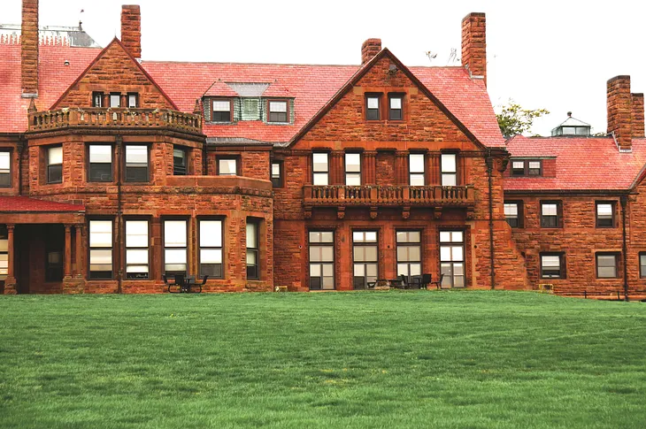 Large estate building with 4 chimneys and 20 windows visible on the two stories. Constructed of brick with two balconies and a large green lawn.