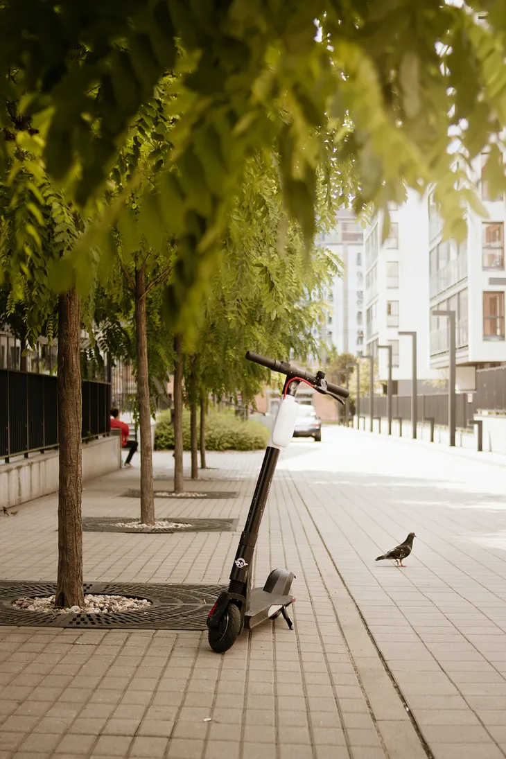A scooter standing up on its kickstand on a public sidewalk with trees.