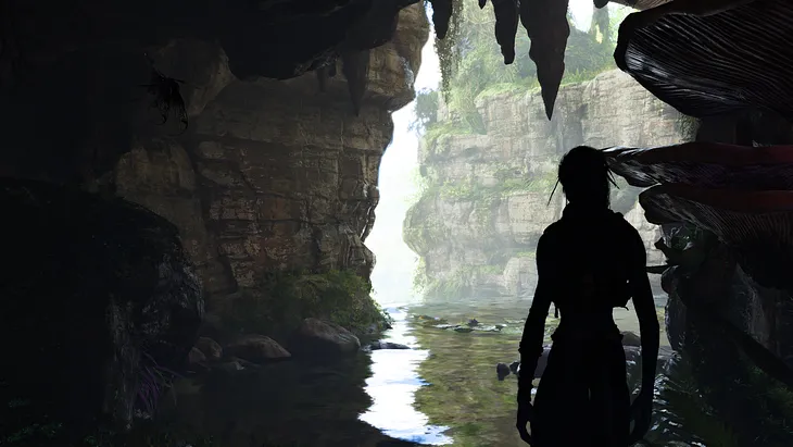 The customizable player character of Avatar: Frontiers of Pandora stands in a cave looking out at a cliff face reflected in a pool.