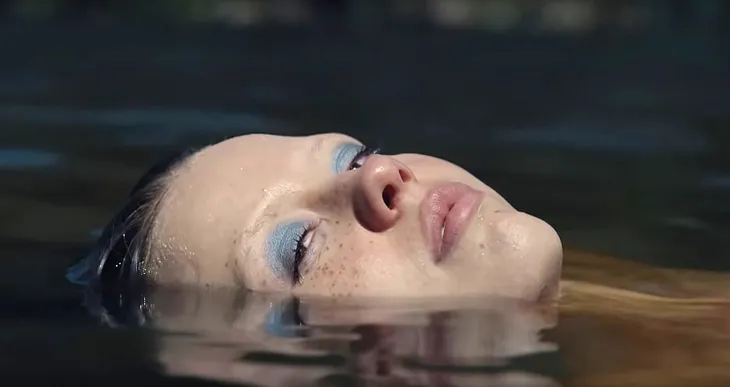 Mia Goth as Maxine is floating in the middle of a pond; only her face is above the water. She wears bright blue eye-shadow and is looking towards the camera through the corners of her eyes.