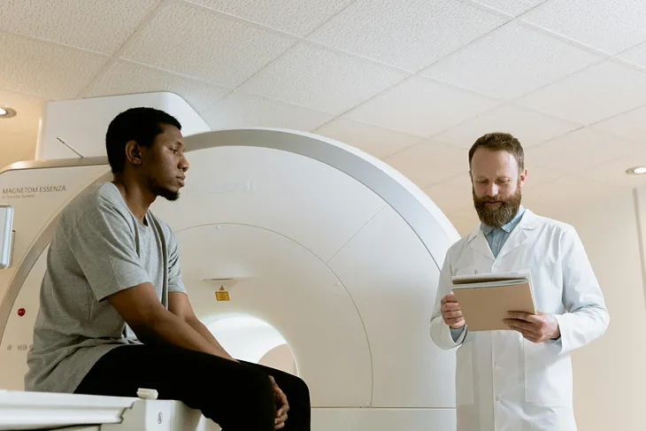 A patient rests on the bed of a CT scan as a doctor looks into their file beside them.